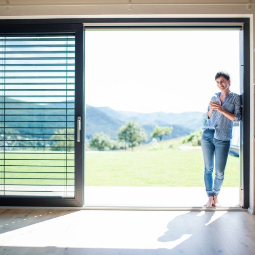 Front view of young woman with coffee standing by patio door at home. Front view of young woman with coffee standing by patio door at home.